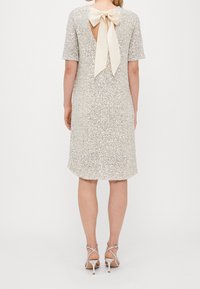 Kortärmad klänning i silver med paljetter, V-rygg och krämfärgad snörning i rosett. Utsvängd silhuett, knä-lång, slät textur.