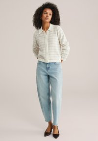 Pletený kardigán v krémovej a béžovej farbe s textúrovaným vzorom, ktorý má zapínanie na gombíky vpredu a špicatý golier. Nosí sa s svetlo modrými džínsami a hnedými lodičkami.