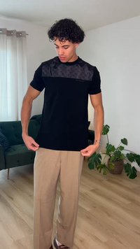 Mann richtet sein schwarzes gemustertes T-Shirt, trägt beige Hose und steht in einem Wohnzimmer mit grünem Sofa und Zimmerpflanzen.