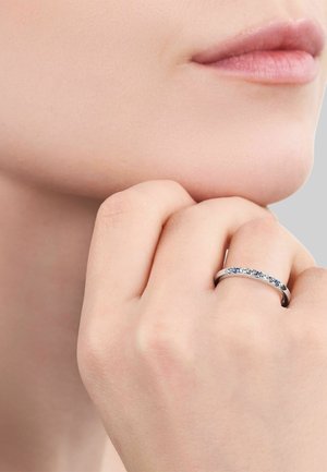 Mano con anillo de plata adornado con pequeñas piedras preciosas azules y transparentes apoyada en la barbilla y los labios de una mujer.