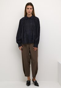 Marineblaues Hemd mit Rüschendetails, kombiniert mit hellbraunen tapered Hosen und schwarzen Absatzschuhen. Glatte Oberfläche, lässige Passform.