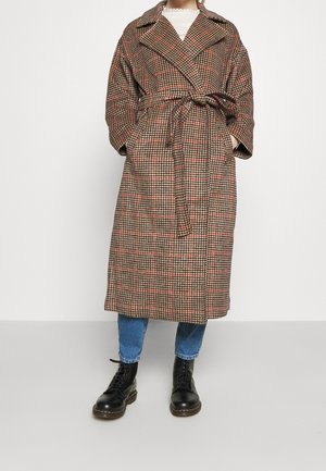 Personne portant un manteau ceinturé à carreaux marron et rouge, un jean bleu, des bottes noires lacées, debout devant un fond blanc uni.