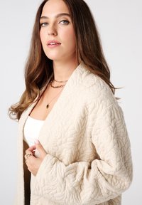 Beige gewatteerde jas met een getextureerd bladenpatroon, open voorkant en brede mouwen, gedragen over een wit shirt, geaccentueerd met gelaagde gouden kettingen.