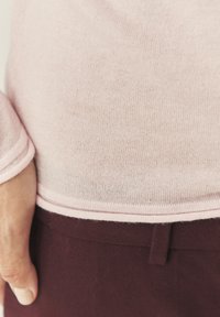Svetloružový pletený sveter s jemnou textúrou, nosený cez tmavé marhuľové nohavice. Má jednoduchý lem s jemným kontrastným stehom.
