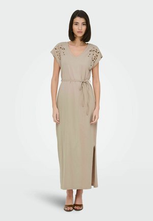 Femme portant une robe maxi beige à manches courtes, découpes aux épaules, fente latérale et ceinture nouée à la taille, debout sur un fond uni.
