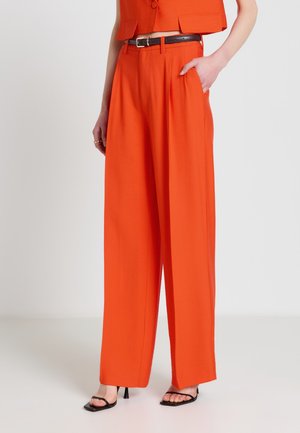 Frau trägt knallorange, hochtaillierte, weit geschnittene Hose mit schwarzem Gürtel und schwarzen offenen Sandalen mit Absatz, Hand in der Tasche.