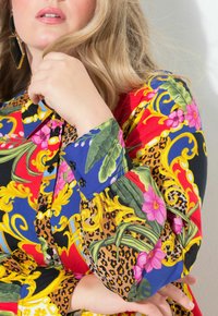 Camisa colorida con una mezcla de rojo, amarillo, azul y negro, con patrones florales y de leopardo; incluye botones negros y un detalle en los puños.