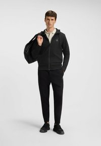 Felpa nera con zip e un design testurizzato, tasche frontali e logo; indossata con pantaloni jogger coordinati e scarpe da ginnastica nere.