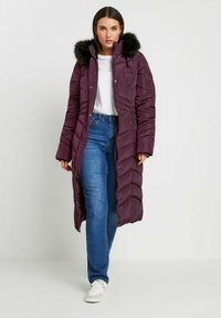 Lilla pufferjakke med hætte af kunstpels, der har et quiltet design, frontknapper og to sidelommer. Båret over jeans og sneakers.