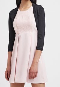 Svart bolero-kardigan över en rosa klänning med en strukturerad spetsöverdel, åtsittande midja och utsvängd kjol med sid sömmar.