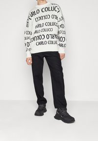 Suéter blanco con texto negro repetido "CARLO COLUCCI", combinado con pantalones negros y zapatillas deportivas negras de suela gruesa. Diseño simple y audaz.