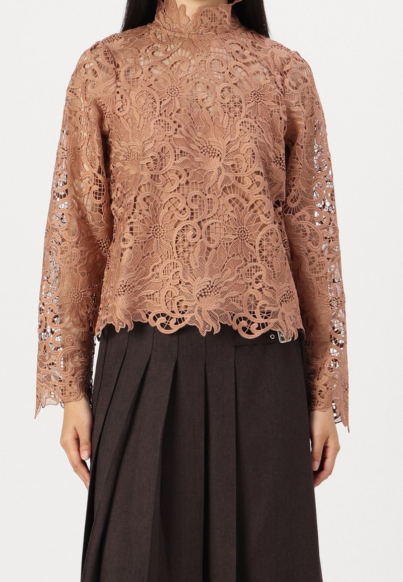 Haut en dentelle couleur rose poudré avec des motifs floraux complexes, col montant et manches longues. Associé à une jupe plissée foncée.