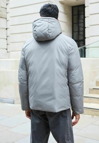 Jachetă puffer gri deschis, cu un guler înalt și o glugă mare. Materialul pare neted și ușor lucios. Purtați peste blugi negri.