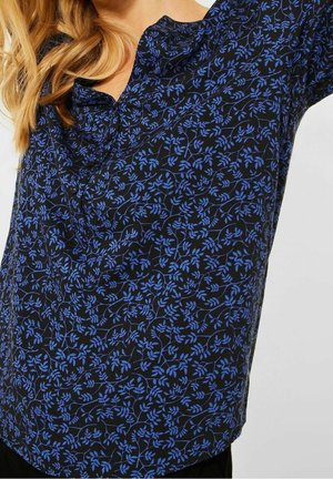 Vrouw draagt een zwarte blouse met een blauw bloemrankenpatroon, knoopsluiting bij de hals en deels zichtbaar blond haar.