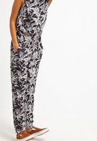 Een zwart-wit jumpsuit met palmbladschema en een strik in de taille, aangevuld met bruine slip-on schoenen en een losse pasvorm.