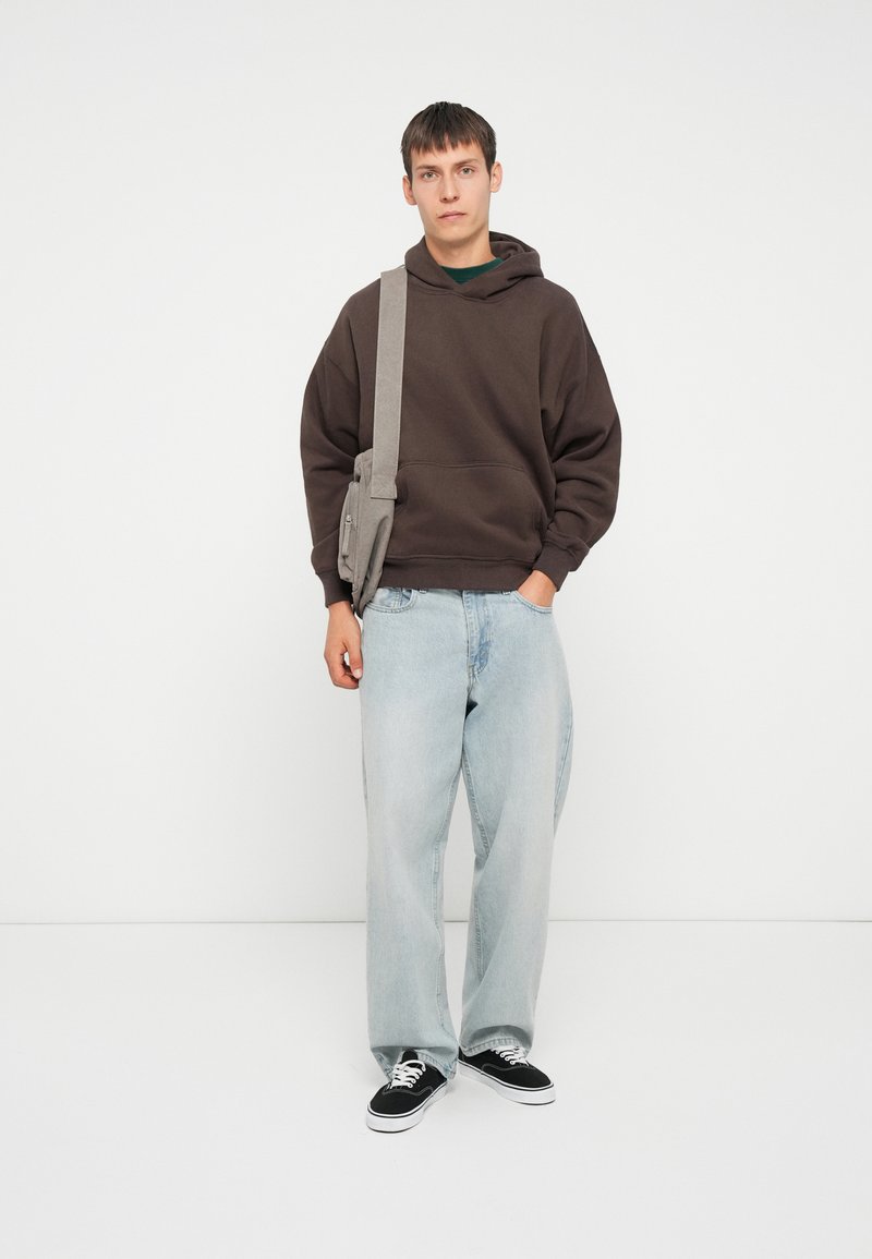 Brauner, übergroßer Hoodie, hellblaue, weite Jeans, schwarze Sneakers. Graue Umhängetasche über die Schulter geschlungen. Schlichtes Design, weiche Texturen.