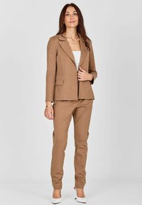 Rjav oblečen suknjič s širšimi črtami, prilagojen blazer in ozke hlače, v kombinaciji z belo bluzo in belimi visokimi petami. Gladka tekstura.