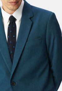 Giacca da abito in lana teal con revers a scalfo, chiusura a un bottone e tasca laterale. Indossata su una camicia bianca e una cravatta nera testurizzata con pois blu.