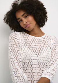 Witte kanten top met bloemenpatroon, ronde halslijn en lange mouwen. Doorzichtige, lichte stof met subtiele textuur en bolle schouders.