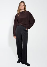 Frau mit langen blonden Haaren, die einen dunkelbraunen lockeren Pullover, schwarze weit geschnittene Hosen und schwarze Schuhe trägt und vor einem weißen Hintergrund steht.