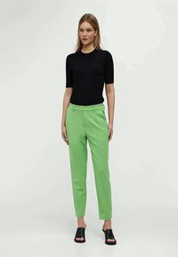 Černý top s krátkým rukávem a zelené kalhoty na míru se slim fit, vyrobené z hladkého materiálu. Kombinováno s černými skluznými sandály.