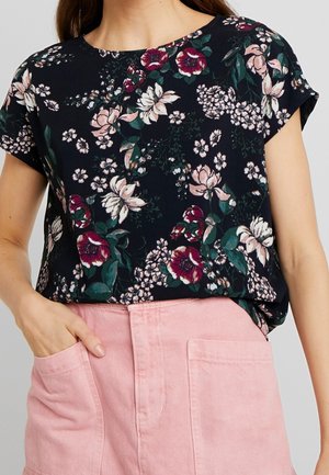 Kobieta ubrana w czarną bluzkę z krótkim rękawem w kwiaty w kolorach różowym, białym i zielonym, do tego jasnoróżowe spodnie z wysokim stanem z kieszeniami.