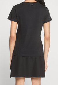 Kobieta w czarnym t-shircie z krótkim rękawem z logo "HUGO" na górze pleców oraz w czarnej spódnicy widocznej z tyłu.