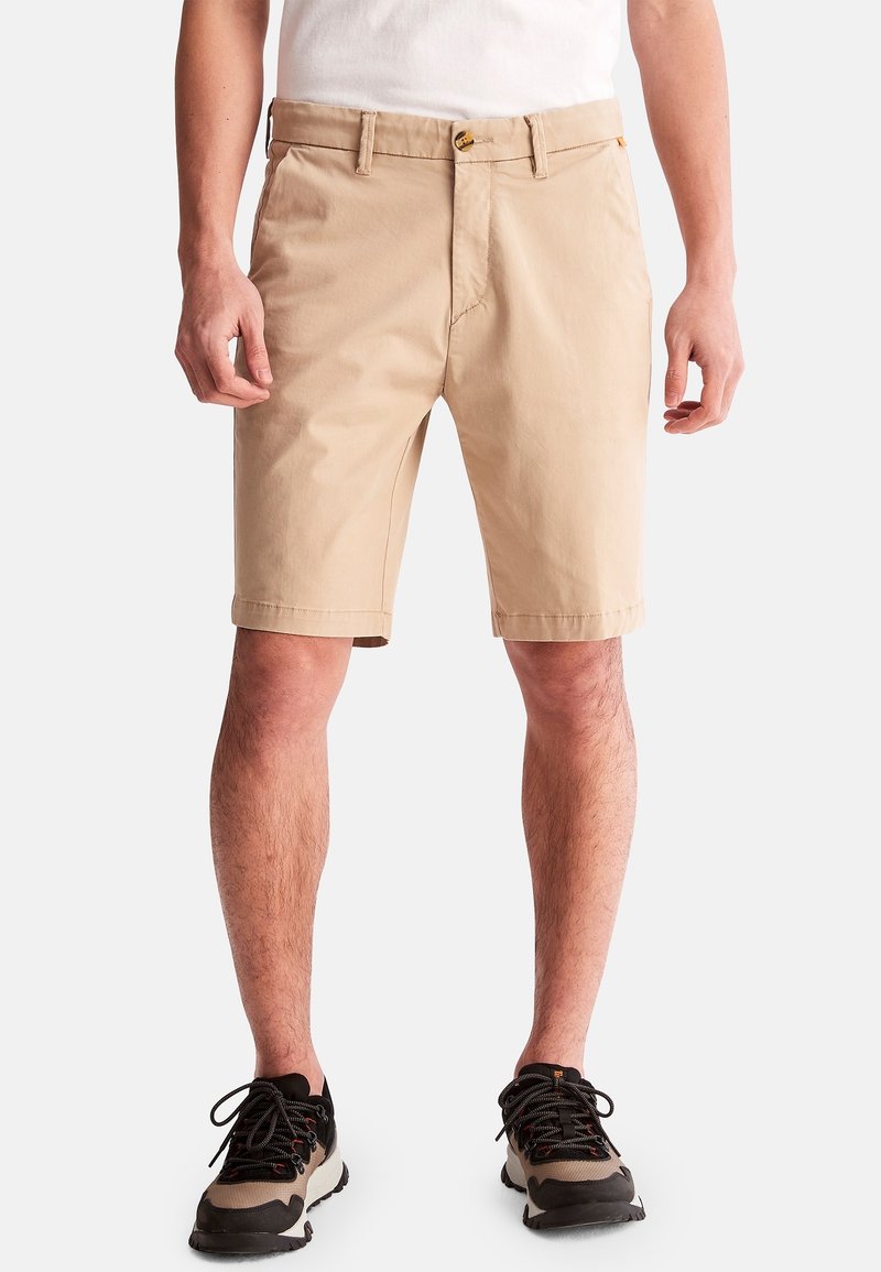 Homme portant un short beige mi-genou, un t-shirt blanc et des baskets noires et beiges, debout devant un fond uni.