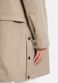 Imperméable beige au toucher lisse, avec une fermeture à boutons sur le côté, des coutures cousues et une coupe légèrement effilée.