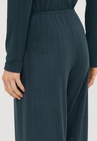 Combinaison côtelée bleu-vert avec des manches longues et une taille cintrée. Tissu doux avec une texture lisse et un éclat subtil, mettant en valeur les détails de couture.