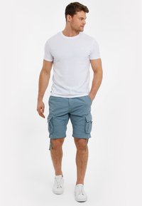 T-shirt bianca a maniche corte abbinata a pantaloni cargo azzurri chiari con tasche laterali e cordini, completata da sneakers bianche.