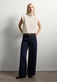 Blonde woman standing with hands in back pockets, wearing a sleeveless beige turtleneck top, dark wide-leg jeans, and brown loafers.
