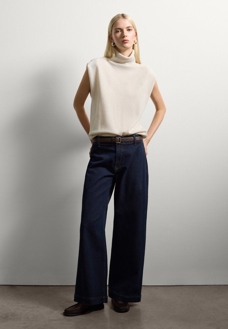 Blonde woman standing with hands in back pockets, wearing a sleeveless beige turtleneck top, dark wide-leg jeans, and brown loafers.