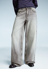 High-waisted, wide-leg gray denim jeans with a faded texture, featuring five pockets and visible stitching, paired with black slip-on shoes.