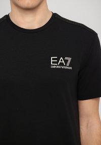 Melnas īsās piedurknes t-krekls ar apaļu kakla izgriezumu, ar baltu EA7 Emporio Armani logo uz krūtīm un strukturētu melnā krāsā detaļu uz pleciem.