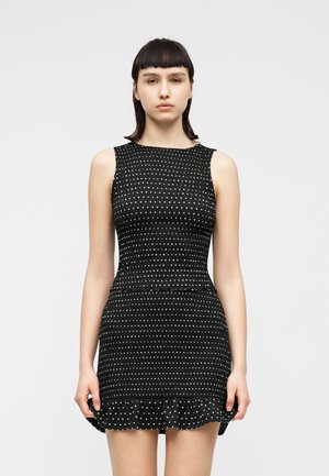 Donna con capelli lisci e scuri che indossa un mini abito nero senza maniche con pois bianchi e orlo arricciato, in piedi davanti a uno sfondo chiaro uniforme.