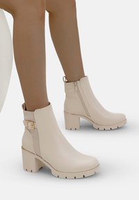 Botines beige con un suave upper sintético, tacón grueso, cremallera lateral y una hebilla decorativa en el tobillo. Suela texturizada para agarre.