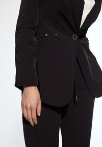 Black blazer with a tailored fit, featuring gold button accents, sleek fabric, and a curved hem. Paired with straight trousers.