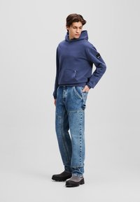 Hoodie azul-marinho com um bolso frontal e etiqueta na lateral, combinado com jeans claros desgastados com costura diagonal e ajuste relaxado. Calçados pretos.