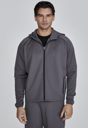 Grå zip-up jacka med huva, gjord av slät tyg. Har en logotyp på bröstet, sidofickor med dragkedja och åtsittande ärmslut. Kombinationen med matchande byxor.