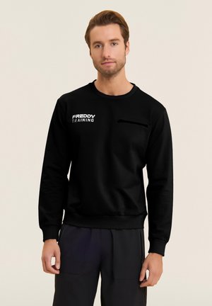 Mies, joka käyttää mustaa collegepuseroa, jossa on "FREDDY TRAINING"-logo, seisoo yksinkertaisen taustan edessä ja katsoo hieman hymyillen kameraan.