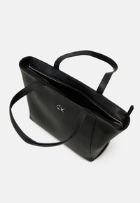 Borsa a tracolla in pelle nera con due manici, chiusura con zip e logo CK argentato sul davanti. Texture liscia e forma strutturata.