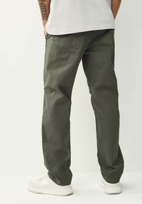 Person wearing loose olive green pants and white sneakers, standing with hands in pockets against a plain light background.