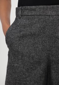 Pantalon à motif pied-de-poule gris, fabriqué dans un tissu texturé, avec une ceinture plate et des poches latérales pratiques.