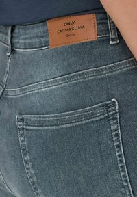 Traperice u srednjem plavom pranju s naglascima u šivanju. Imaju kožnu oznaku označenu "ONLY CARMKOMA" blizu struka.