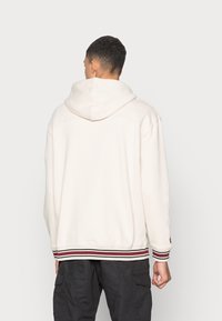 Crème hoodie met ribbelzoom met rode en witte strepen, gemaakt van zachte stof, met een relaxte pasvorm en verlaagde schouders.