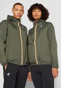 Giubbotti leggeri verdi con cerniere gialle e bianche, dotati di cappucci regolabili, indossati sopra pantaloni neri, con logo sui giubbotti.