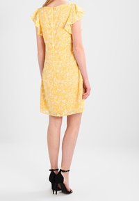 Kvinna klädd i en kort gul blommig klänning med volangärmar och svarta högklackade skor med ankelsnodd, stående mot en vit bakgrund.