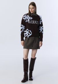 Vrouw in een donkere trui met de tekst "VARIOUS" en bloemenpatronen, geruite rok en donkerbruin kniehoog schoenen, staand op een effen achtergrond.