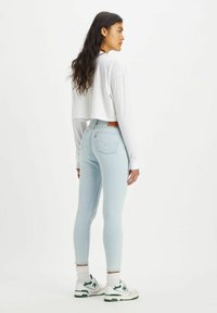 Chemise blanche à manches longues et cropped, jeans skinny bleu clair, et sneakers blanches avec des accents verts. Logo et étiquette Levi's visibles à la taille.